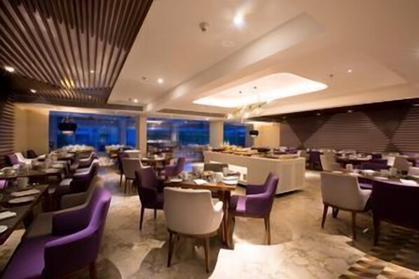 A modern hotel dining area with multiple tables and chairs, a central buffet, and a view of the cityscape. - Le Méridien Goa, Calangute (Calangute)
