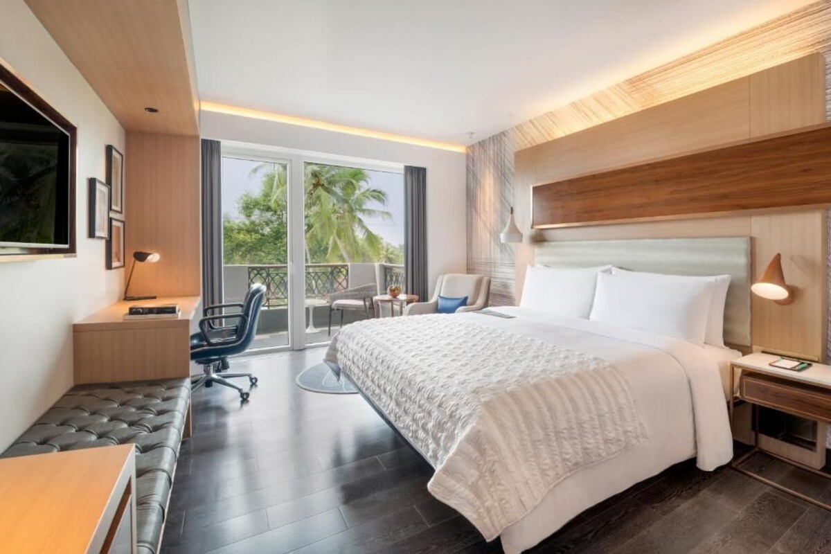 A modern hotel room with a large bed, a desk, a chair, and a balcony with a view of palm trees.