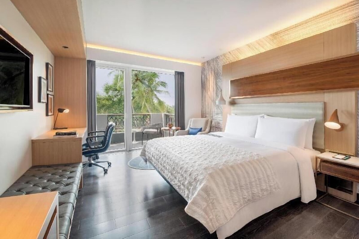 A modern hotel room with a large bed, a desk, a chair, and a balcony with a view of palm trees.