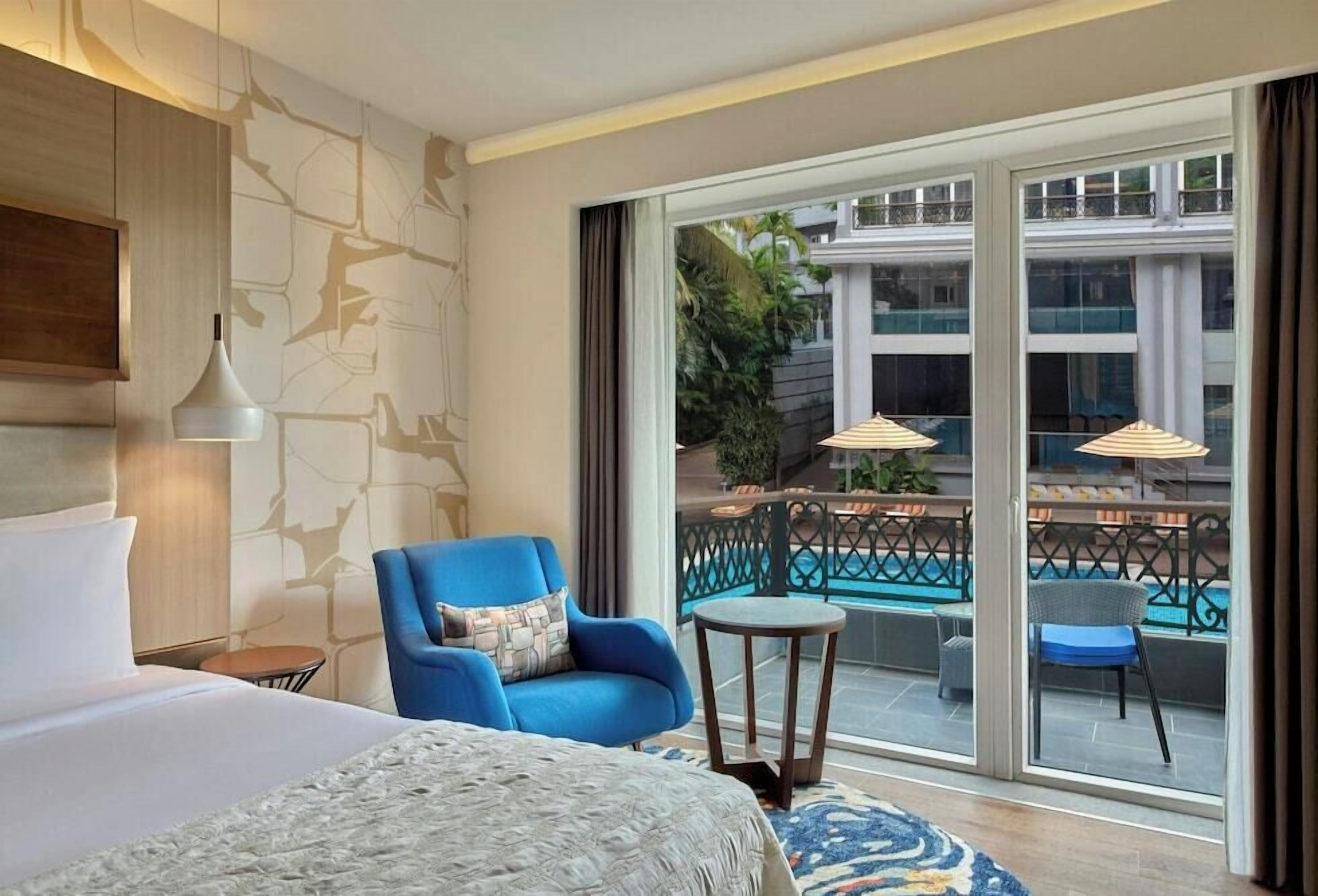 A hotel room with a bed, a blue armchair, a small round table, and a view of a swimming pool through large windows.