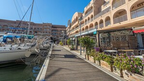 A marina with boats docked, a wooden walkway, and buildings with balconies and shops.