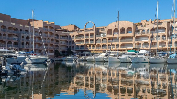 A marina with multiple boats docked, a large building with balconies, and clear blue skies.