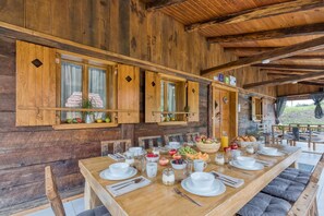 A wooden dining table set for breakfast with various fruits and beverages, wooden walls, and a view of the outdoors.