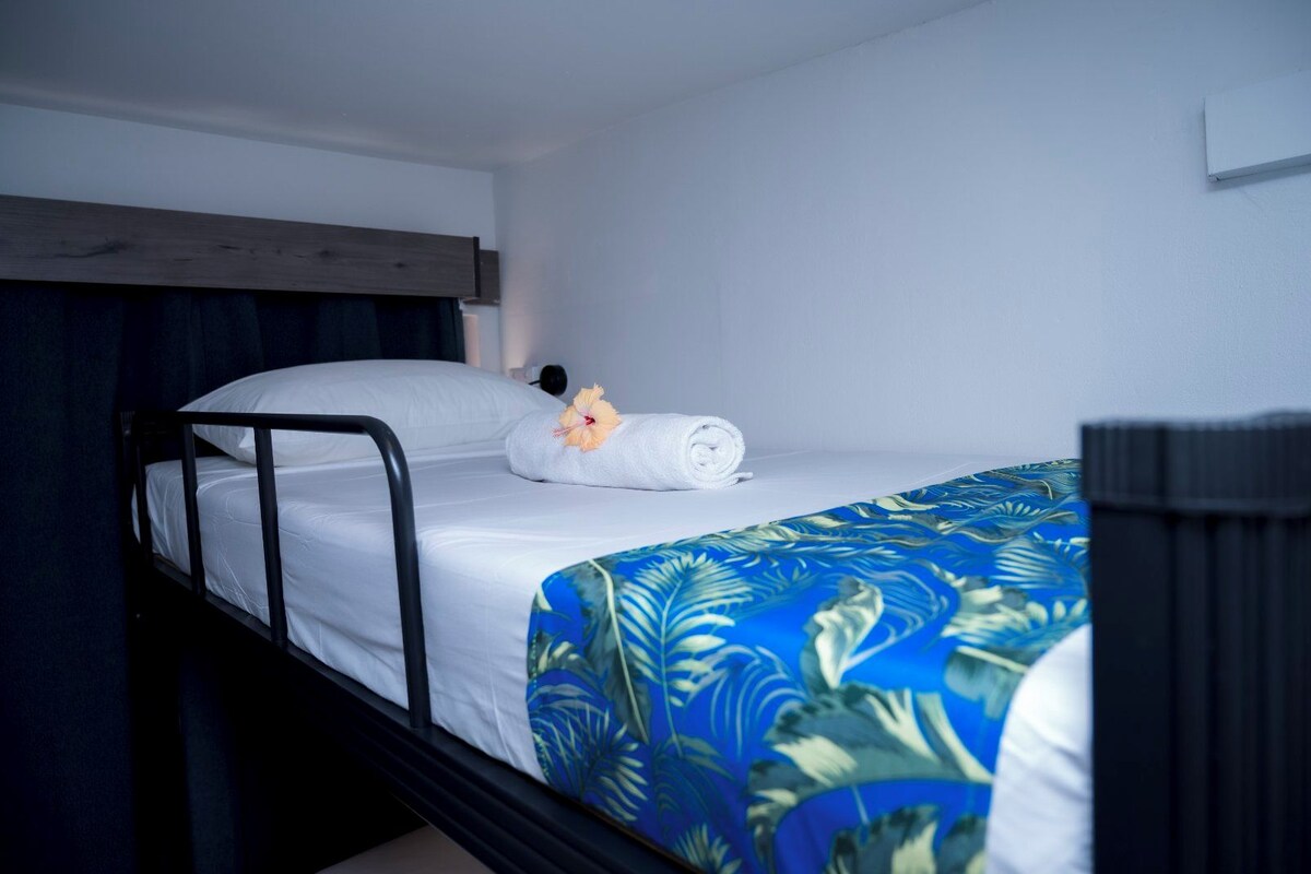 A single bed with a white sheet, a rolled towel, and a hibiscus flower.