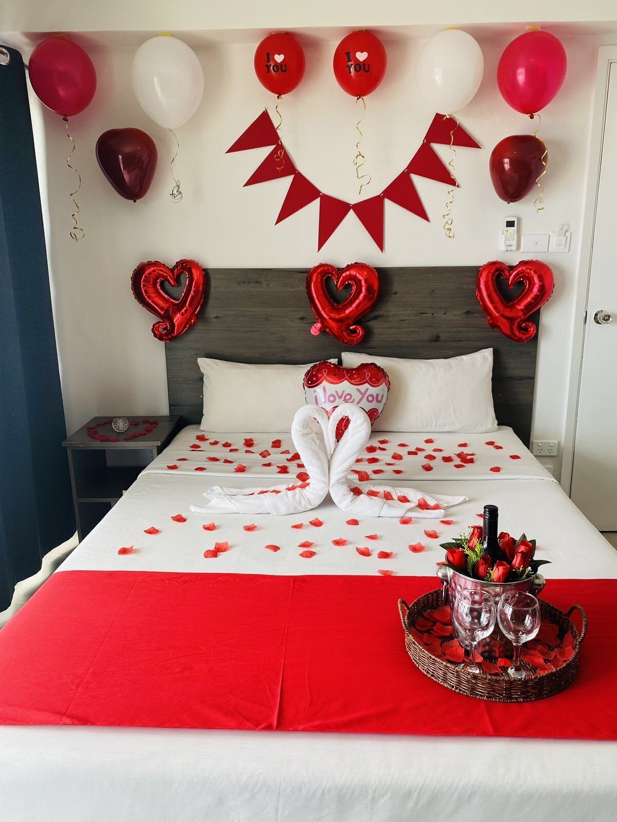 A hotel room with a bed adorned with red and white decorations, including a heart-shaped towel arrangement and balloons with "I love you" messages.