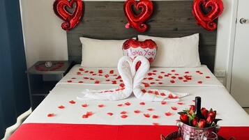 A hotel room with a bed adorned with red and white decorations, including a heart-shaped towel arrangement and balloons with "I love you" messages.