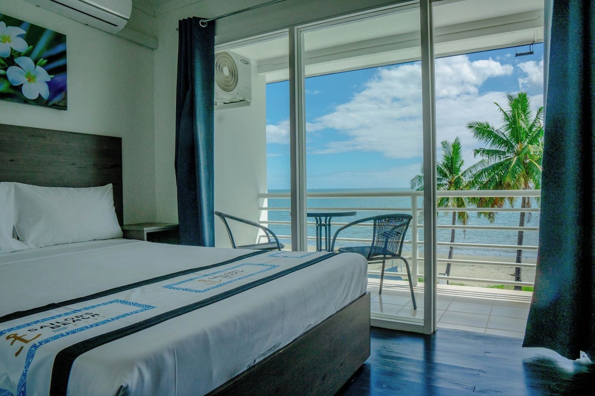 Habitación de hotel con cama, vistas al océano, balcón con mesa y sillas, y una planta tropical afuera.