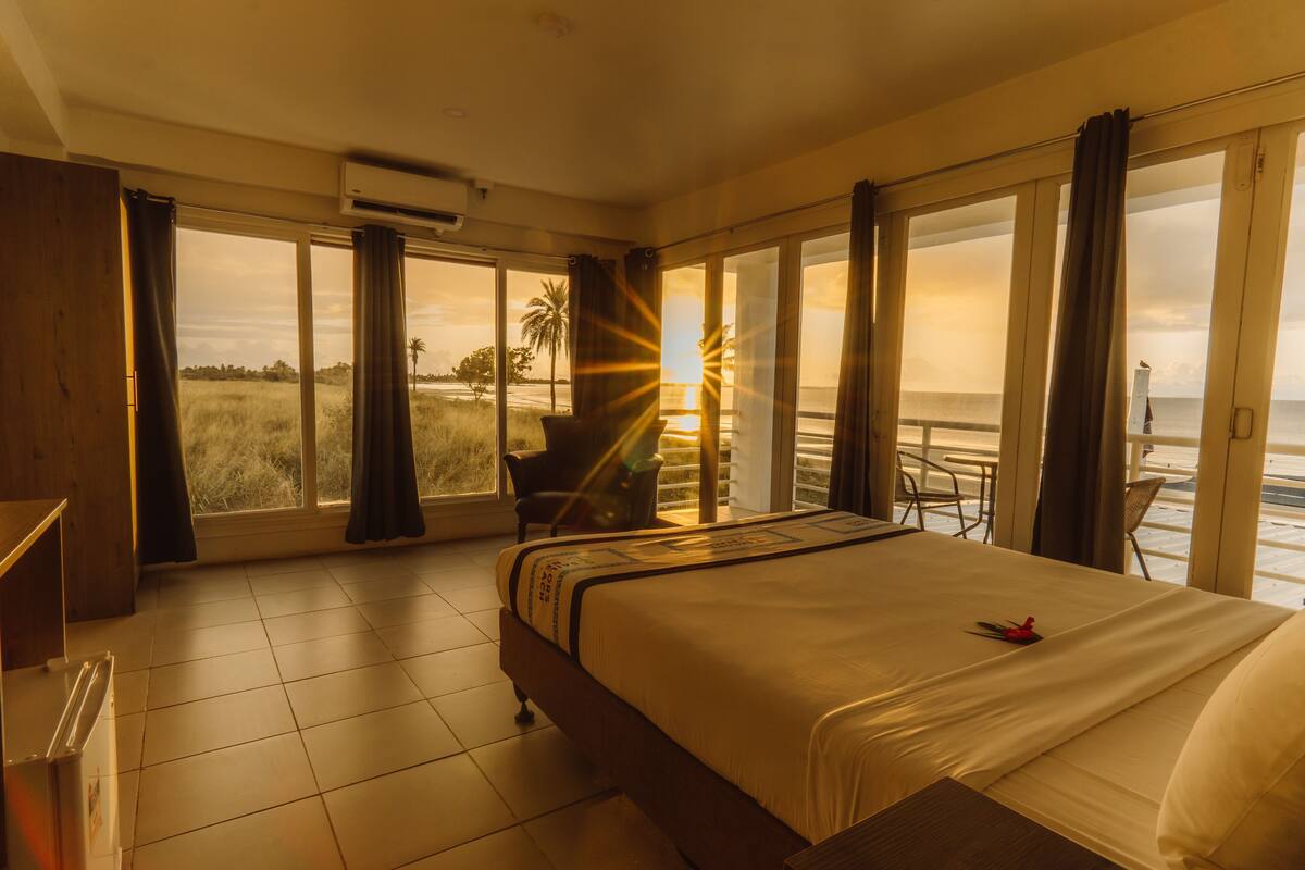 A hotel room with a large bed, a chair, a table, and a view of the beach and sunset through large windows.