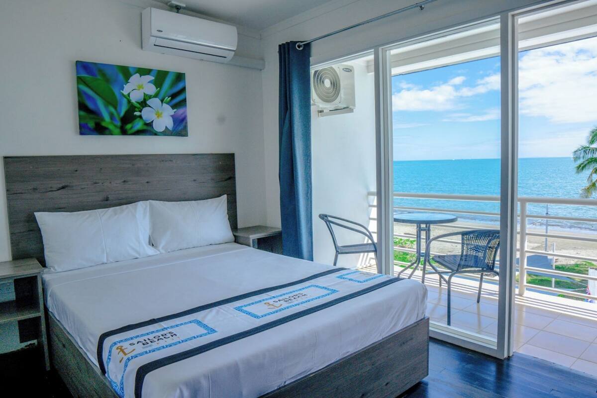A hotel room with a bed, a painting of flowers, an air conditioning unit, and a balcony with a view of the sea.