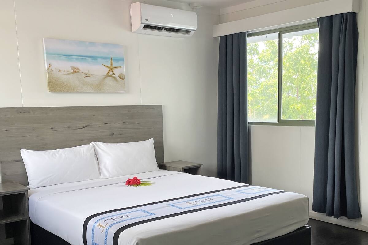A hotel room with a large bed, a nightstand, a wall-mounted air conditioner, a window with curtains, and a beach-themed painting on the wall.