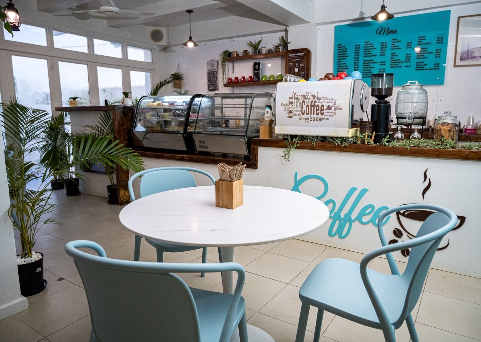A coffee shop interior with a round table, blue chairs, a menu board, and a coffee machine.