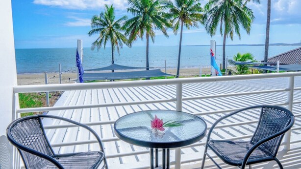 A balcony with two chairs and a table, overlooking a beach with palm trees and a flag. - Sailors Beach Fiji (Nadi)