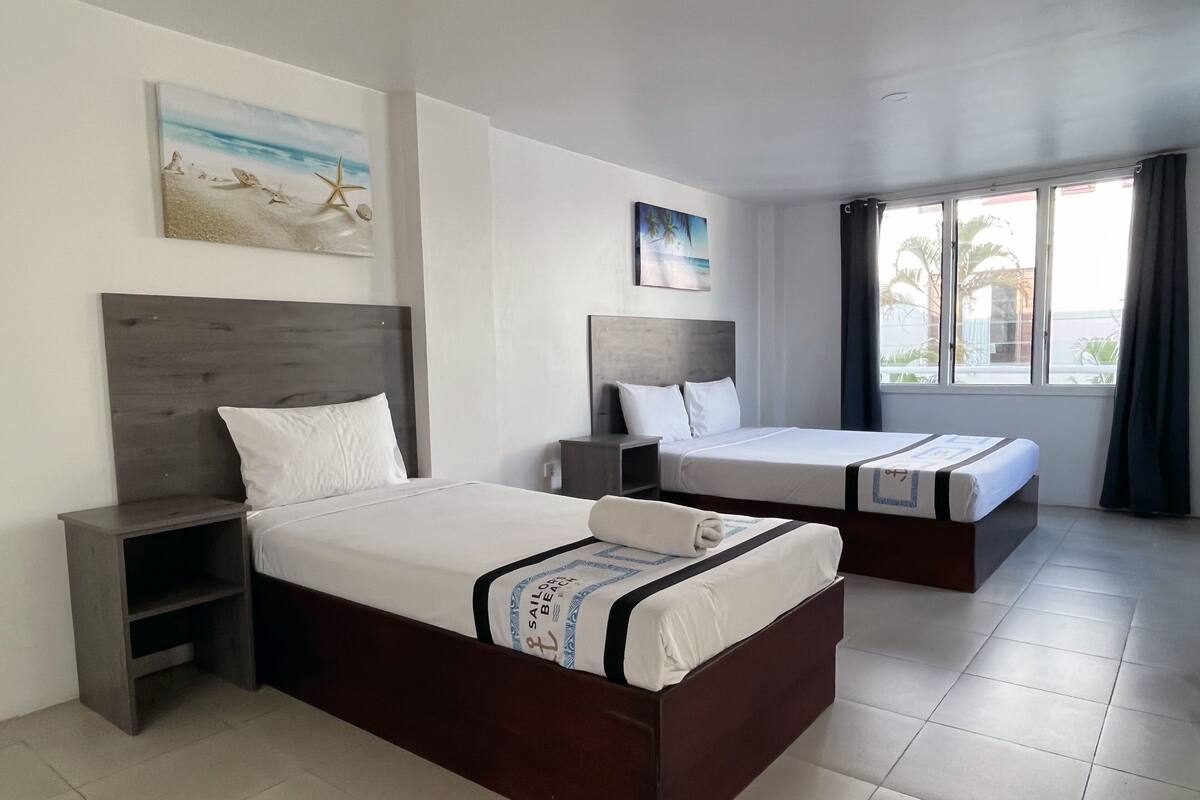 A hotel room with two beds, a nightstand, a window with curtains, and artwork on the walls.