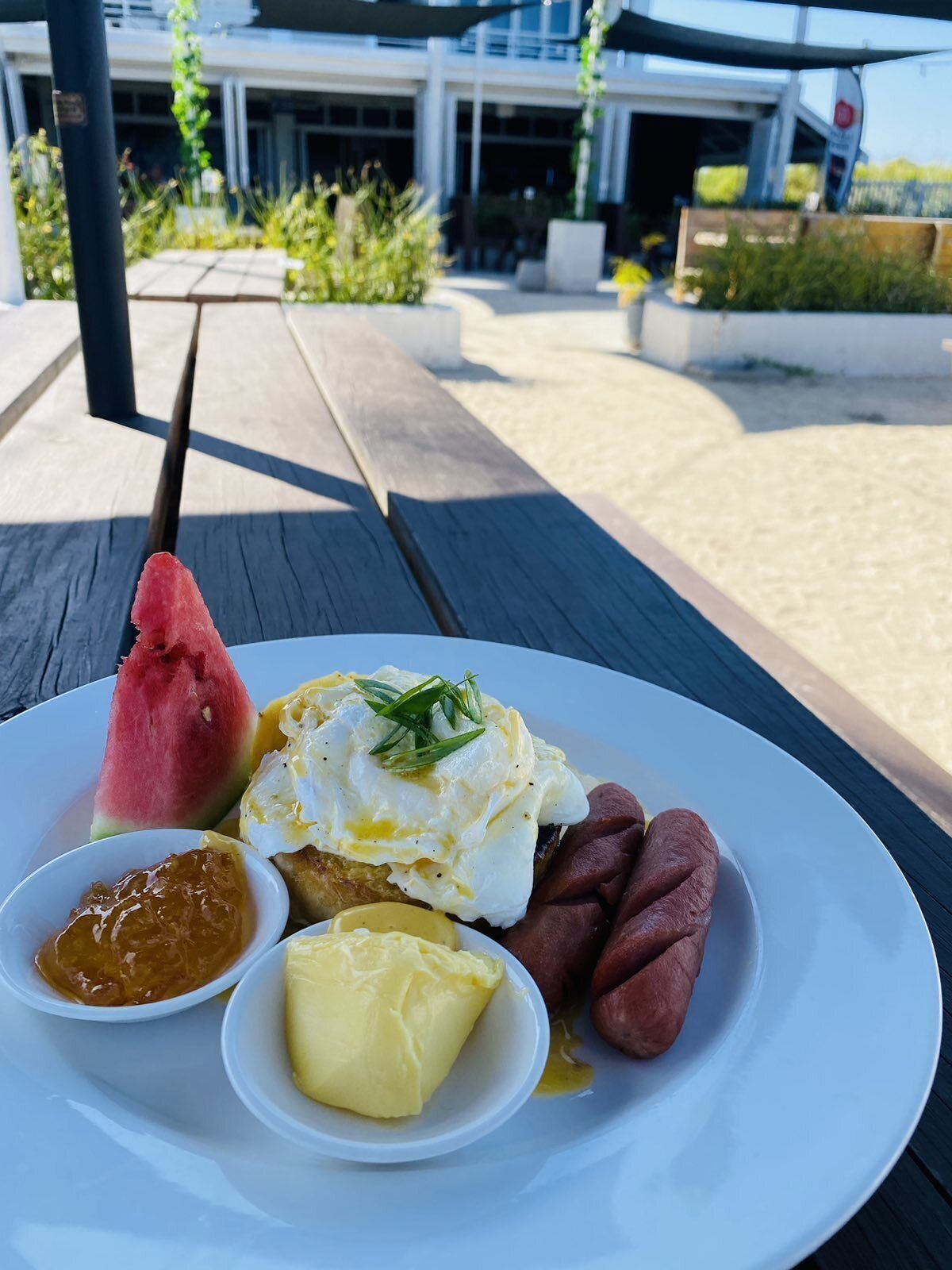 Un plato de comida con brochetas de sandía, huevos pochados, salchichas y condimentos sobre una mesa de madera.