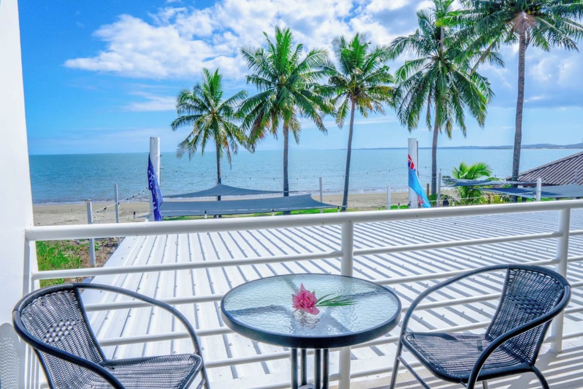 A balcony with two chairs and a table, overlooking a beach with palm trees and a flag.