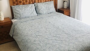 A bed with a patterned bedspread, a wooden headboard, and two pillows. There is a painting on the wall above the bed and a nightstand with a lamp to the left.