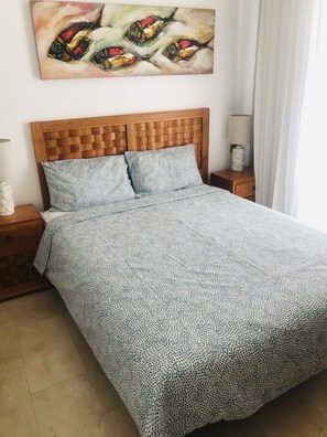 A bed with a patterned bedspread, a wooden headboard, and two pillows. There is a painting on the wall above the bed and a nightstand with a lamp to the left.