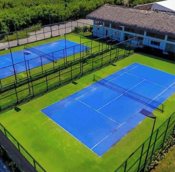 A tennis court with a net, surrounded by a green fence, and a building with a thatched roof in the background.