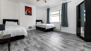 A modern bedroom with two beds, a ceiling fan, a large window with curtains, and a black wardrobe. - House - Sleeps 10 - Garden - Close to City Centre (Colchester)