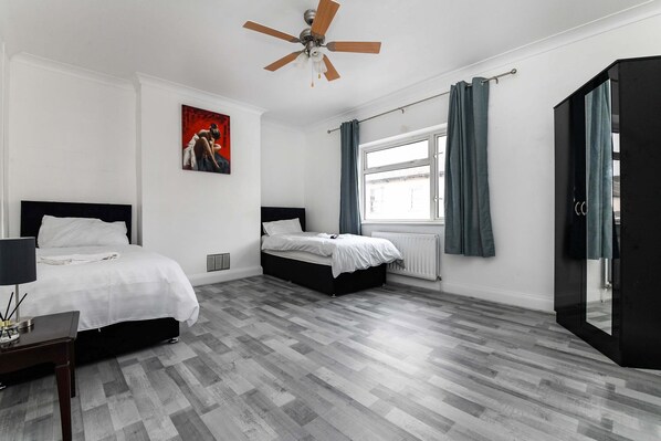 A modern bedroom with two beds, a ceiling fan, a large window with curtains, and a black wardrobe. - House - Sleeps 10 - Garden - Close to City Centre (Colchester)