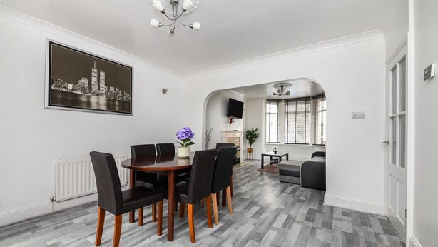 A modern dining area with a wooden table and chairs, a cityscape artwork on the wall, and a view into a living room with a sofa and a TV. - House - Sleeps 10 - Garden - Close to City Centre (Colchester)