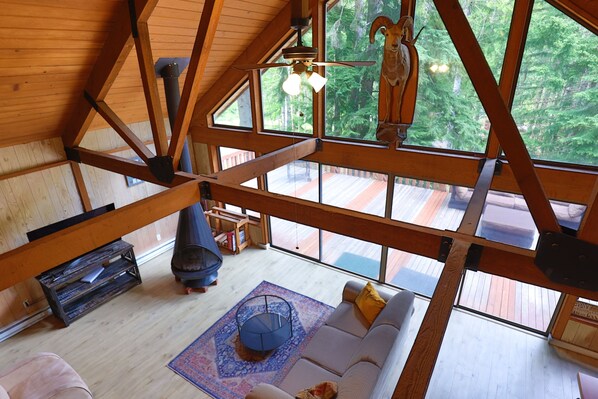 Una habitación con techos altos y vigas de madera, un sofá blanco, una silla azul y un amplio ventanal con vistas a un bosque.