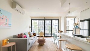 Ein modernes Wohnzimmer mit einem grauen Sofa, einem runden Hocker und einem Essbereich mit Barhocker. Ein großes Fenster bietet einen Blick auf Gebäude im Freien.