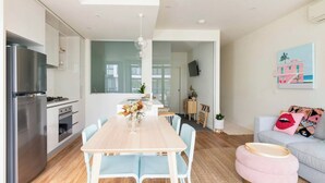 Eine moderne Küche mit einer großen Kochinsel, einem Essbereich mit langem Tisch und Stühlen sowie einem Wohnbereich mit Sofa und Kissen.