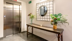 Een lift met een bordje waarop staat dat deze niet gebruikt mag worden bij brand, een wandspiegel en een houten console tafel met twee planten in potten.