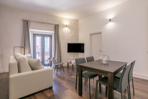 A modern living room with a sofa, dining table, chairs, and a flat-screen TV mounted on the wall. - Sierpes Luxury Premium de 2 -1 bedroom (Sevilla)