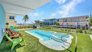 A swimming pool with a hot tub, surrounded by green lawn and colorful chairs, with a view of a waterfront and buildings in the background. - Dock, Pool, FREE RAMP, Kayaks, Jacuzzi, Amazing Ocean Views Pets are Welcomed! (Marathon)