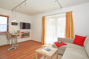 Un salon moderne avec un plancher en bois, un canapé gris agrémenté d’un coussin rouge, une petite table avec des magazines, un téléviseur fixé au mur et un coin repas avec une chaise et une table.