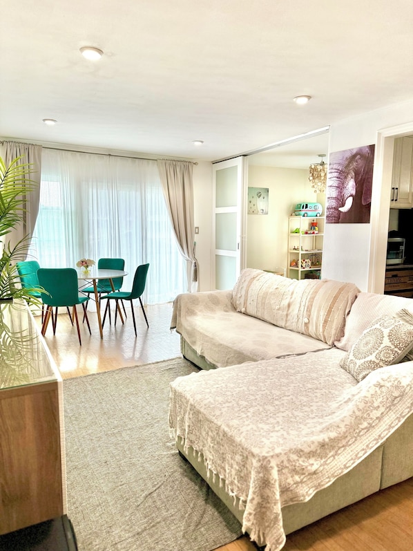 Un salon avec un canapé, une table à manger avec des chaises, un coin cuisine et une grande fenêtre avec des rideaux.