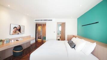 A modern hotel room with a large bed, a wooden headboard, a teal accent wall, a small desk with a plant, and a framed photo on the wall.