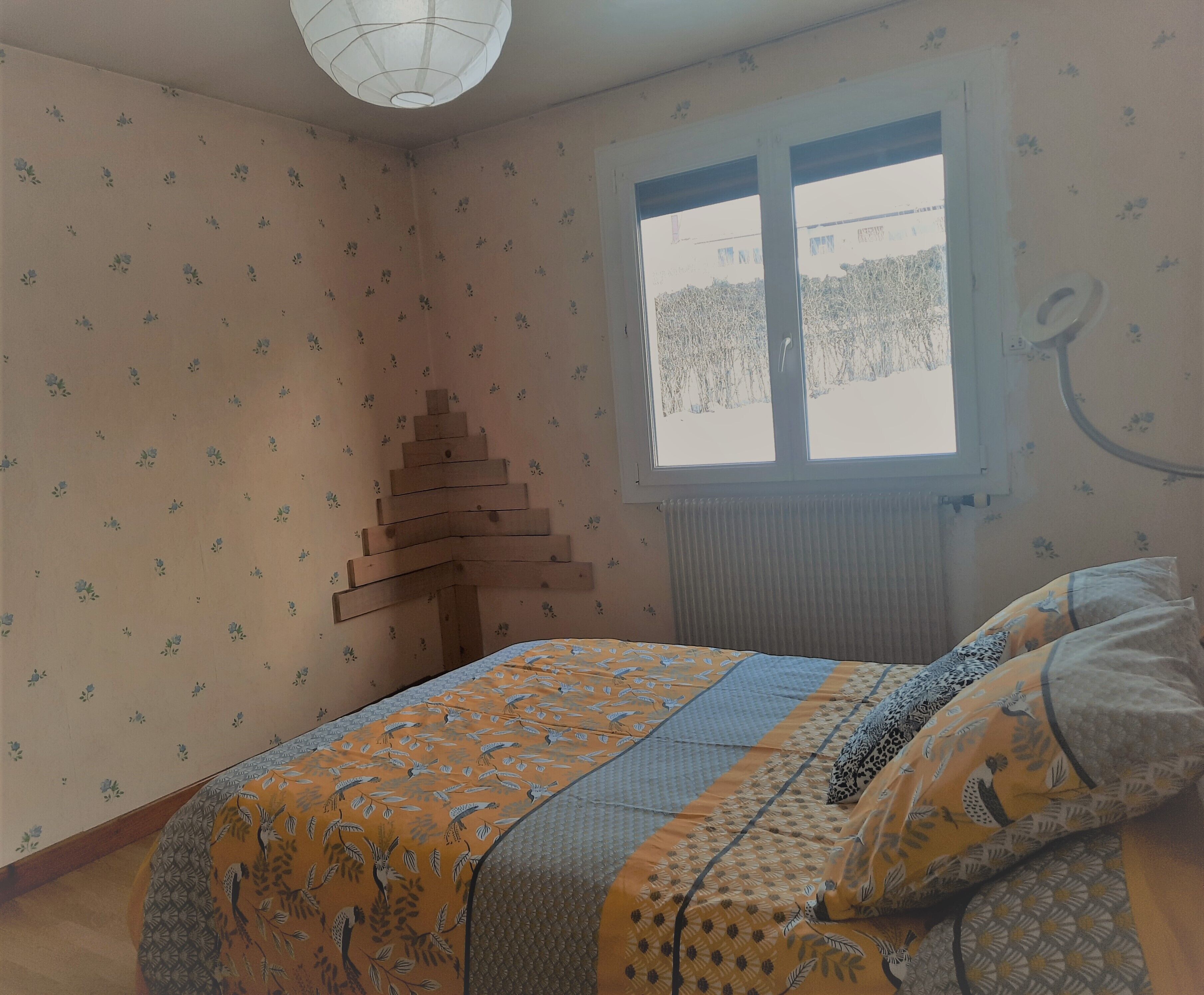 A bedroom with floral wallpaper, a bed with patterned bedding, a window with a view, and a unique wooden structure on the wall.