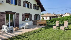 Una casa de dos pisos con contraventanas de madera, un patio con mobiliario exterior y un césped bien cuidado.