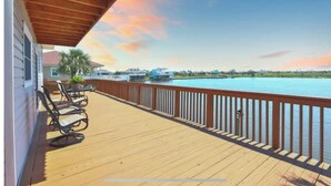 A wooden deck with a railing overlooking a body of water, a boat, and a distant shoreline. - Waterfront Terramar Getaway, Amazing Sunsets, Outdoor Bar, Kayak, Beach, Fishing (Galveston)
