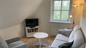 A modern living room with a white sofa, a small round table, a bookshelf with books, a TV, and a window with a view of trees.