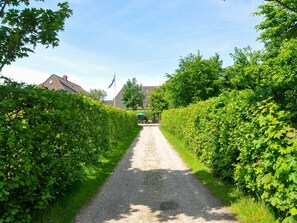 Un chemin de gravier bordé de haies verdoyantes et luxuriantes, menant à un bâtiment sur lequel flotte un drapeau.