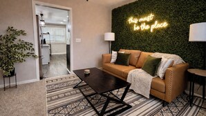 Un salon avec un canapé marron, un tapis à motifs, un ventilateur de plafond et un mur vert orné d’un néon.