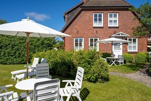 Ein Backsteinhaus mit einem gepflasterten Weg, der zu einer Terrasse mit weißer Gartenmöbeln und Sonnenschirmen führt.