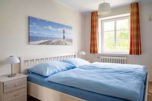 Una camera da letto con un letto, un comodino, una lampada, una finestra con tende e un quadro raffigurante un faro appeso al muro.