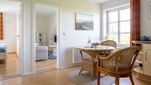 A room with a dining table, wicker chairs, a view of a mountain through a window, and a white cabinet with a blue pot. - Apartment Oland - House on the dike (Utersum)