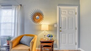 A room with a beige armchair, a small wooden table with a lamp, a decorative mirror, and a white door.