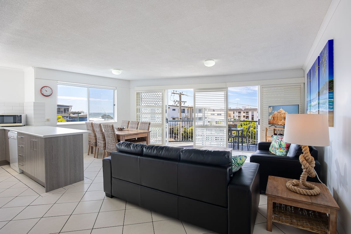 Eine moderne Küche mit Kochinsel, ein schwarzes Ledersofa, ein Essbereich mit Stühlen und Blick durch große Fenster.