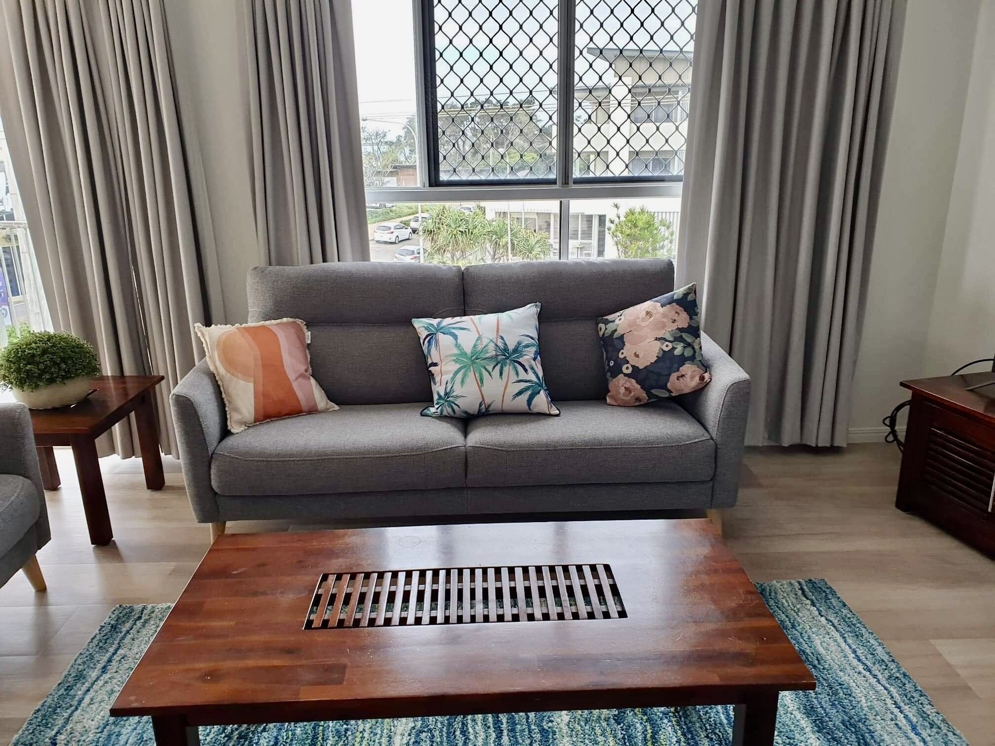 A modern living room with a grey sofa, three decorative pillows, a wooden coffee table with a vent, and a view of the street through a window with a metal grid.