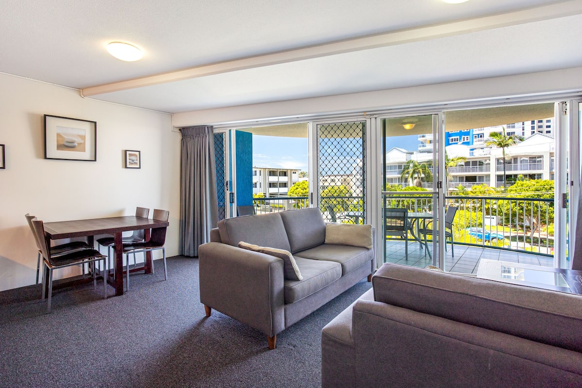 Ein modernes Wohnzimmer mit grauem Sofa, einem Esstisch mit Stühlen und einem Balkon mit Blick auf Gebäude und Grünflächen.