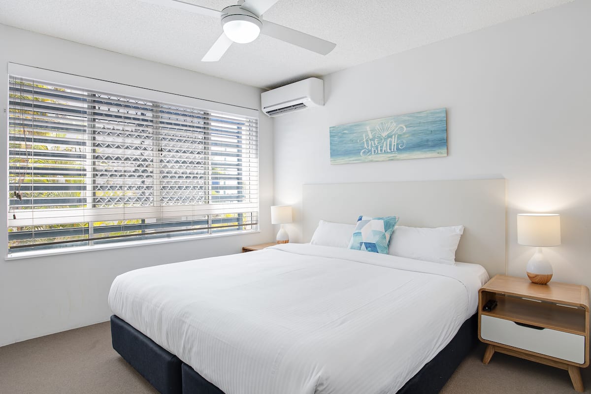 Ein modernes Schlafzimmer mit einem großen Bett, Deckenventilator, Klimaanlage, Nachttisch mit Lampe und einem Fenster mit Jalousien.