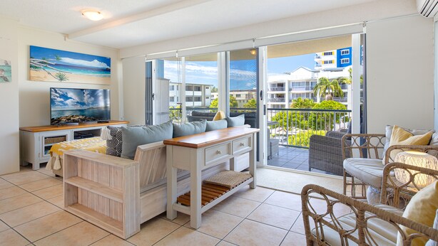A spacious living room with a large TV, comfortable seating, and a balcony with a view of the city. - Coral Sea Apartments (Maroochydore)