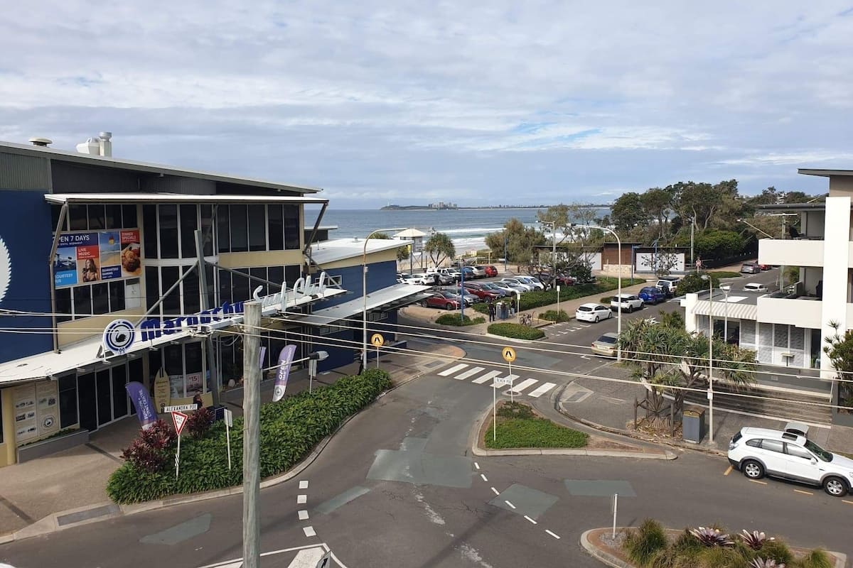 Blick auf eine Küstenstraße mit einem Gebäude mit blauer Fassade und mehreren Bannern, einem Parkplatz mit Autos und freiem Blick auf das Meer.